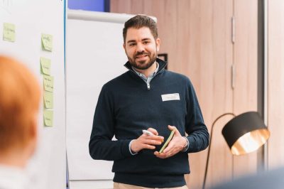Mann hält Schulung vor Flipchart, trägt dunklen Pullover, beige Hose; helles Zimmer, warme Lampenbeleuchtung.